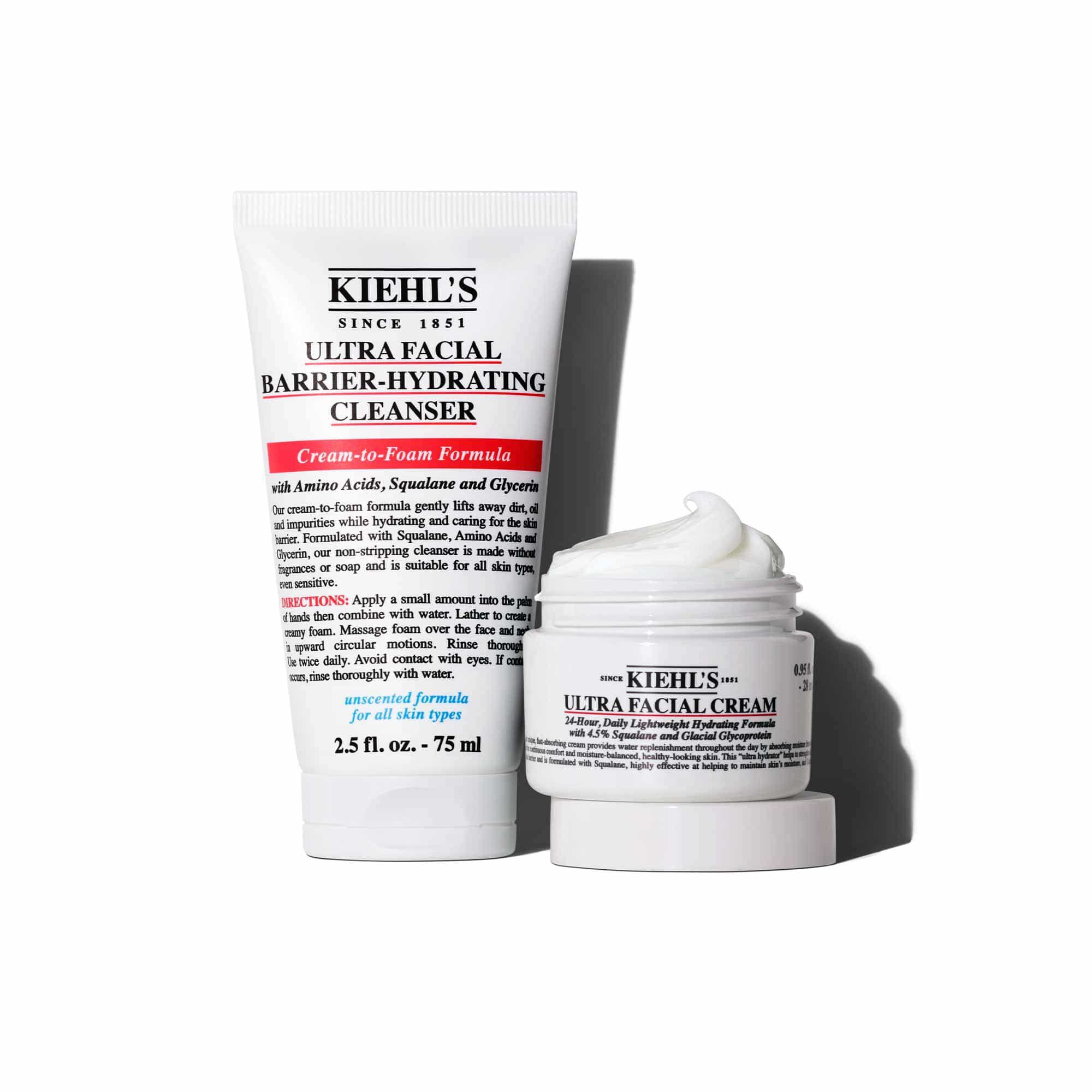 A graphic displays two Kiehl's products: a white tube of "KIEHL'S ULTRA FACIAL BARRIER-HYDRATING CLEANSER”, standing upright next to an open white jar of "KIEHL'S ULTRA FACIAL CREAM". The jar's lid is placed under it and the entire set is on a white background.