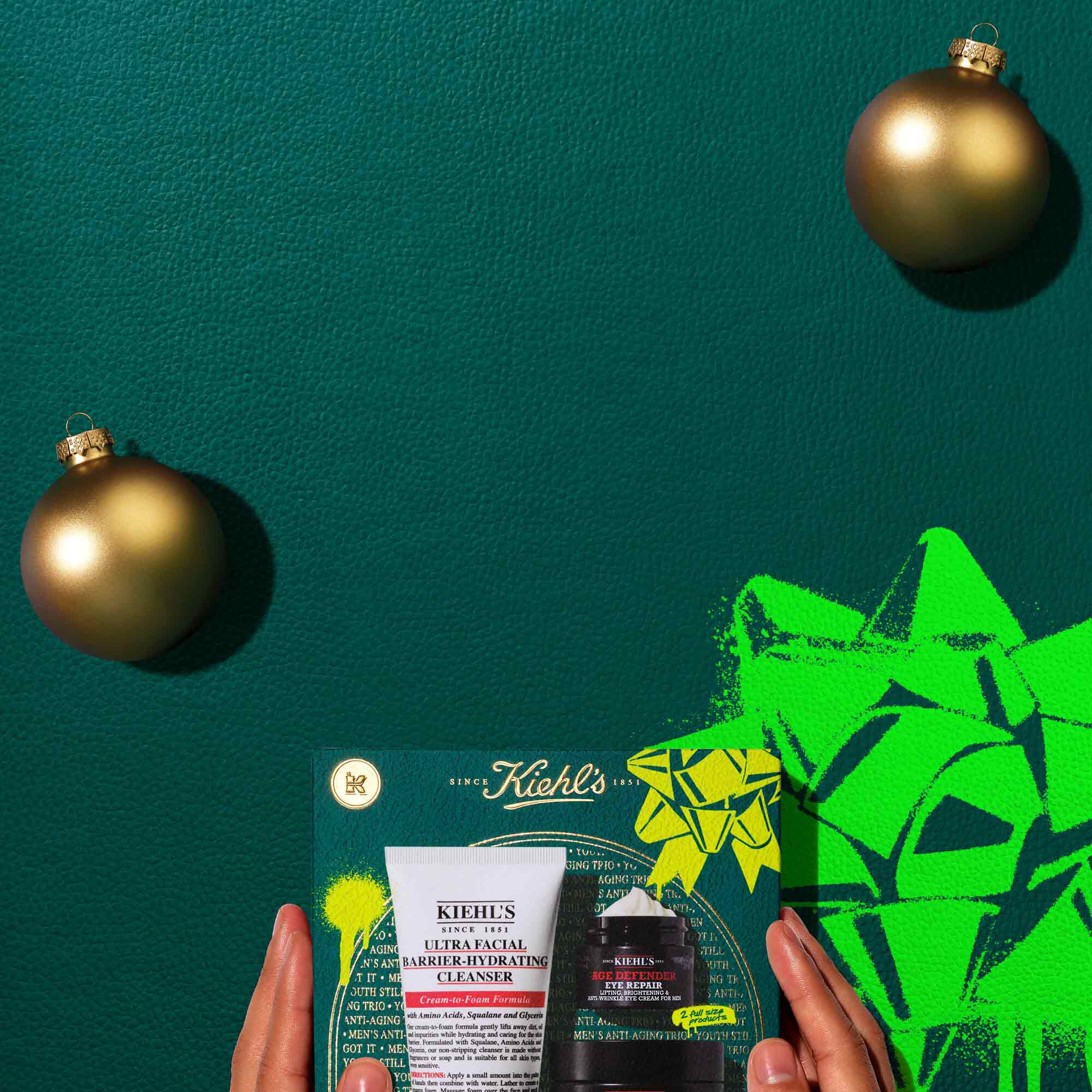 A graphic, set against a dark green textured background with a bright green graphic bow on the lower right, features an arrangement of products and a gift set. At the top left is a white tube of "KIEHL’S AGE DEFENDER EXFOLIATING CLEANSER." Next to it, a small black jar of "KIEHL’S AGE DEFENDER EYE REPAIR" and a larger black jar of "KIEHL’S AGE DEFENDER MOISTURIZER" are displayed. Two gold spherical ornaments accent the scene. Below, a Kiehl’s gift box displays the same three products. The gift box is decorated with bright green graphic shapes and a bow design.